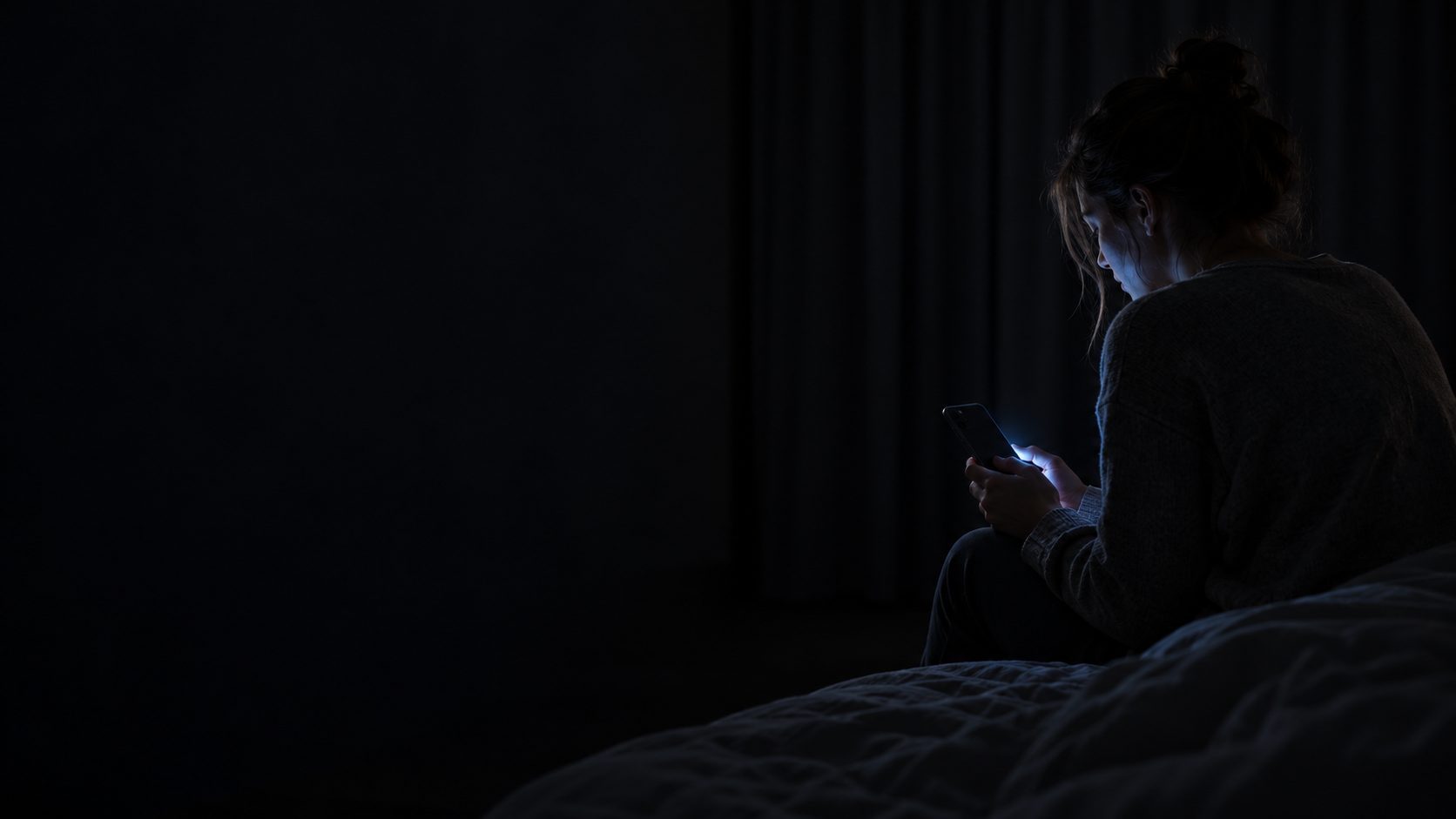 A person sits on the edge of a bed at night, lit only by a phone screen.